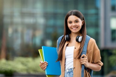 Neşeli öğrenci güneşli kampüste kitap tutuyor. Canlı kampüste kitapları kucaklayan mutlu genç bir kadın pozitiflik ve gençlik dolu akademik ruhlar yayıyor sırt çantası taşıyor, kopyalama alanı yaratıyor.