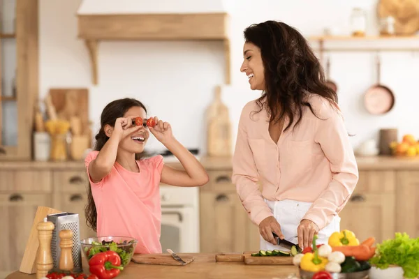 Joyful mom and daughter girl enjoying cooking time together while preparing vegetable salad indoor, kid peeking through pepper slices as eyeglasses in modern kitchen. Fun and healthy eating