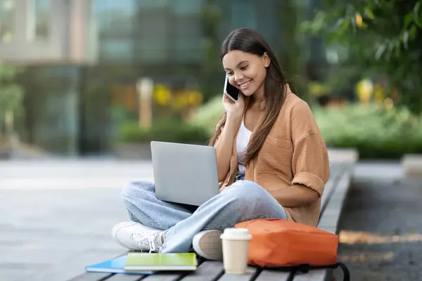 一个快乐的女学生在一个空荡荡的大学校园里打电话 在笔记本电脑上打字 很可能在网上讨论功课 或者协调一个学习小组 抄袭空间 — 图库照片