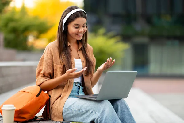Smart spirited young woman student interacts with her laptop and headphones, perhaps web surfing for academic articles or collaborating on a group project, have video call, university campus