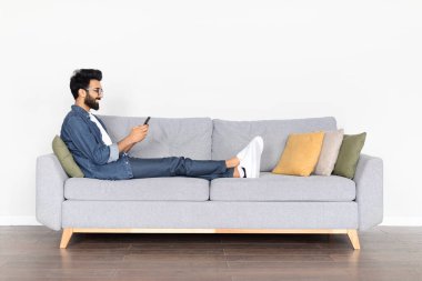 Genç Hintli bir adam, gri bir kanepede rahatça uzanan, akıllı telefonuyla meşgul olan, beyaz bir arka planda izole edilmiş, rahatlama ve dijital yaşam tarzını canlandıran,