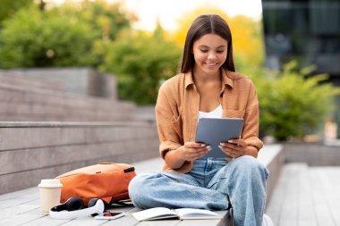 Neşeli genç bayan öğrenci dijital tabletinde ışıltılı bir gülümseme ile geziniyor, dingin, yeşil üniversite kampüsünde rahatça oturuyor, barışçıl bir akademik ara veriyor, güzel mobil uygulama.
