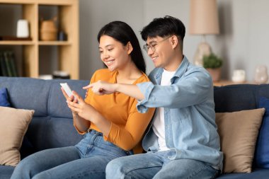 Neşeli Asyalı çift internet içeriğini akıllı telefondan paylaşıyor, evdeki bir kanepede rahatça oturuyorlar, eşleri boş zamanlarında ve teknoloji kullanımında. Telefon başında yeni uygulama gösteren koca noktaları