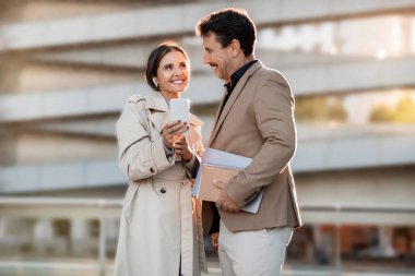 Kendini işine adamış iki iş arkadaşı, orta yaşlı bir adam ve kadın, telefon ekranını paylaşırken iş görüşmesi yapıyorlar, bir şehir sokağında duruyorlar, işbirlikçi ruhu sergiliyorlar.