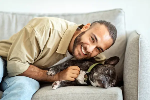 Best Friend Concept. Portrait of happy millennial man petting his cute french bulldog. Affection And Love For Animal. Guy lying on the couch and having fun together with his pet in living room