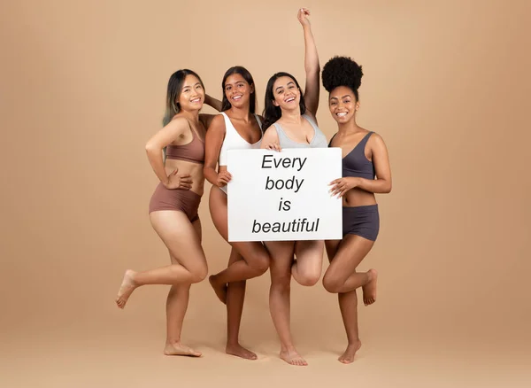 Four multiracial women confidently pose in studio, holding placard with the empowering message Every body is beautiful, celebrating all body types, full length