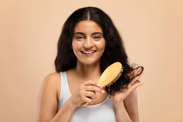 Beauty Routine. Attractive happy indian woman brushing her long curly hair with bamboo comb, beautiful smiling eastern female using wooden brush standing over beige studio background, copy space