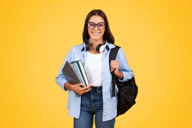Sırt çantalı, kitaplı, gözlüklü ve kulaklıklı genç bayan öğrenmenin tadını çıkarın, sarı stüdyo arka planında izole edin. Okulda eğitim, başarı, yaşam tarzı ve proje