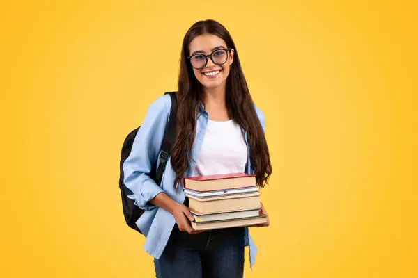戴眼镜背着背包的快乐的女学生拿着很多书 喜欢学习 在黄色的工作室背景下独处 大学教育 生活方式 家庭作业和图书馆 — 图库照片