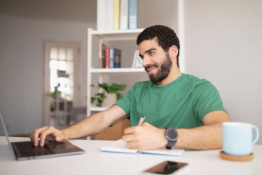 Neşeli genç Arap blogcu, menajer, iş yerinde dizüstü bilgisayarla yazı yazan, uzaktan çalışan, ofisin iç kısımlarında notlar alan. Proje, girişim, iş ve öğrenim