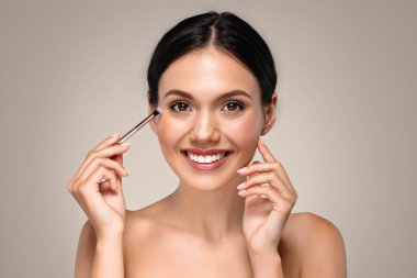 Mutlu milenyum Avrupalı bayan makyajını küçük fırçayla yapıyor, doğal mükemmel cilt bakımından hoşlanıyor, gri stüdyo arka planında izole edilmiş. Temel makyaj, yaşam tarzı, rutin prosedür