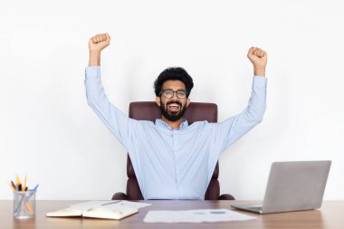 Hint milenyum adamı profesyonel, pozitiflik ve başarı yayıyor, ellerini kaldırıyor, laptopuyla etkileşime geçiyor, masada oturuyor, online başarı sahnesini özetliyor.