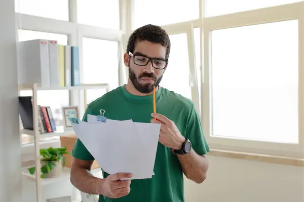 緑色のTシャツとメガネのペンハンサムな男が文書を保持し 光のオフィスのインテリアで鉛筆で熟考しました アイデア スタートアップソリューション ブレインストーミングの作成 — ストック写真