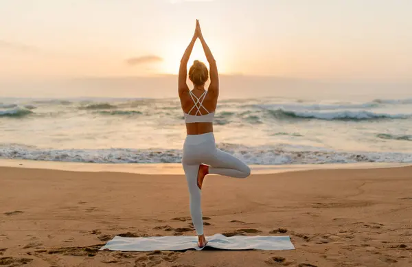 Woman in white activewear strikes a balanced yoga pose, with the gentle sunset illuminating the waves and the sandy beach behind her, back view, full length