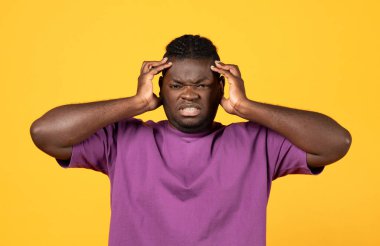 Kafasına dokunan, rahatsızlık ve acı veren, sarı arka planda şiddetli baş ağrısı ya da migren semptomlarıyla başa çıkan siyahi genç adam, şakaklara, tıbbi durumlara ya da sağlık sorunlarına elini bastırıyor.