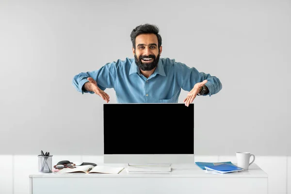 Happy indian businessman pointing at computer monitor with blank black screen at office, mockup. People, technologies and business concept