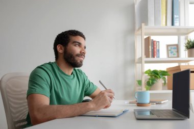 Bin yıllık düşünceli Arap öğrenci ya da menajer notlar alıyor, dizüstü bilgisayarda planlama günü, aydınlatma odasının iç mekanında fikir yaratıyor. Uzaktan iş kurmak, okumak, online ders, iş, yaşam tarzı