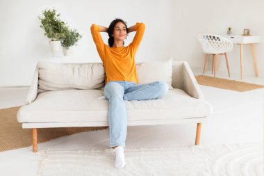 Keyifli bir gülümsemeyle kanepede dinlenen, modern oturma odasında mola veren, evdeki boş zamanlarını kutlayan sıradan bir Arap kadın. Bir anlık sakinliği kucaklamak.