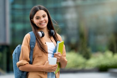 Elinde kitaplarla gülümseyen bir öğrenci, üniversite kampüsünde poz veriyor. Kitapları tutan, üniversitede duran, neşe ve bilimsel uğraşların bir karışımını sembolize eden parlak, genç bir kadın.