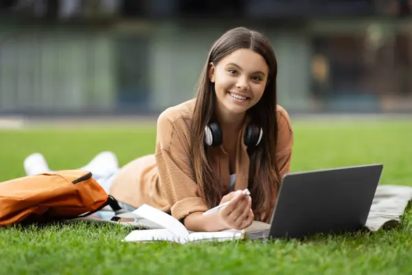 在空荡荡的大学校园的草坪上 一位精力充沛 快乐的年轻女性正在与笔记本电脑打交道 可能是打电话或参加在线讲座 做笔记 对着摄像机微笑 — 图库照片
