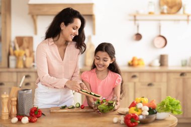 Neşeli anne ve kız çocuk, modern mutfak içlerindeki taze organik sebzelerden salata yapıp, kesim malzemeleri ekleyerek eğleniyorlar. Sağlıklı beslenme alışkanlıkları, aile beslenmesi.