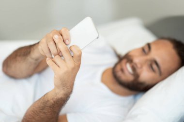 Cep telefonu kullanan, interneti tarayan ve telefonda mesaj atan evde yatağında yatan gülen adam. Sosyal medyada Smartphone Networking ile Mutlu Milenyum Adamı. Yeni Mobil Uygulama Ters Konsepti