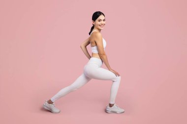 Beyaz spor giyimli enerjik ve atletik kadın dinamik bir hamle pozisyonu benimsiyor. Sağlığını ve zindeliğini tam uzunlukta, soluk pembe arka plana karşı vurguluyor.