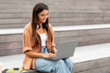 Kampüsteki bankta tek başına oturan neşeli genç bayan öğrenci, dizüstü bilgisayarında parşömenler ve yazılar, muhtemelen sakin ortamda yapılacak sınavlar için araştırma ya da çalışma,