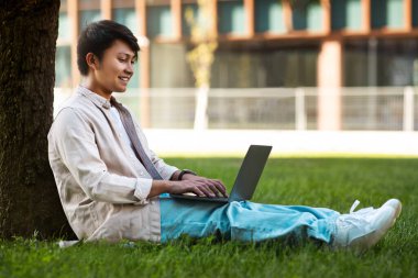 Ağacın altında oturan konsantre Asyalı adam dışarıda, parkta güzel bir atmosferde dizüstü bilgisayarıyla çalışıyor. Teknoloji, iletişim, eğitim ve uzaktan çalışma konsepti, kopyalama alanı