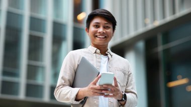 Sıradan ve CEO 'su olan yakışıklı Asyalı genç adam ofise gidiyor, elinde dizüstü bilgisayarla yürüyor, sokak sokak yürüyor, telefon kullanıyor, e-postaları kontrol ediyor, iş telefonu uygulaması kullanıyor, gülümsüyor, boşluk kopyalıyor.