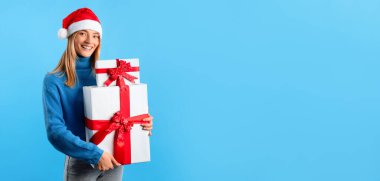 Noel, yeni yıl indirimi. Noel Baba şapkalı mutlu bayan elinde hediye kutuları ve mavi stüdyo arka planında kameraya gülümsüyor. Panorama, boş yer. Noel kutlaması, kış tatili havası.