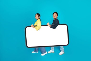 Mavi stüdyo arka planında izole edilmiş, boş ekranlı, büyük akıllı telefonlu neşeli Asyalı çift. Online alışveriş uygulaması, reklam ve teklif, sohbet, cihaz ile yaşam tarzı