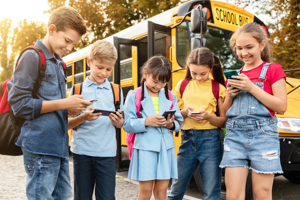Gadget addiction in children. Diverse preteen kids using smartphones while standing near yellow school bus outdoors, multiethnic boys and girls playing mobile games, scrolling, browsing internet