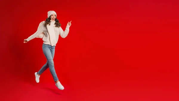 伟大的圣诞礼物 兴奋的年轻女子在圣诞老人帽和针织物跑来跑去跳过红色工作室的背景 放眼空旷的空间寻找文字 广告圣诞销售 — 图库照片