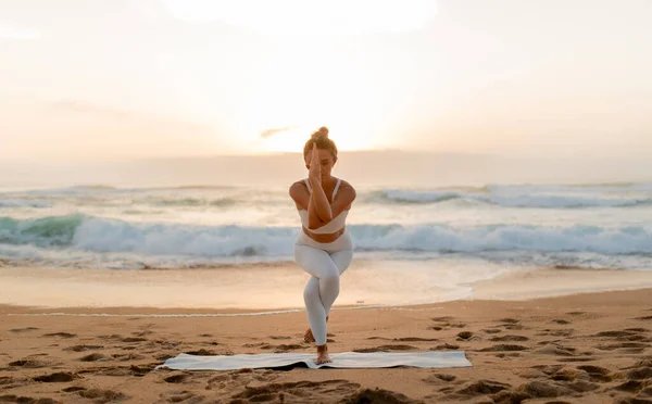 白いアクティブウェアの女性は 彼女がヨガのポーズを保持するように深く焦点を当て 日没は彼女の背後にある衝突波と砂浜の上に暖かい光を投げます — ストック写真