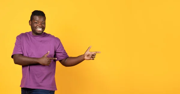 看那里 快乐的年轻黑人男人在空旷的空间边指路 快乐的非洲裔美国人男人指示广告设计的自由位置 站在黄色工作室的背景上 — 图库照片