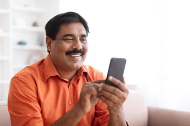 Ev Rahatlaması. Olgun Hintli adam akıllı telefonu kontrol ediyor. Orta yaşlı bir adam cep telefonunu dikkatle karıştırıyor, rahat bir oturma odasında oturuyor, boşluğu kopyalıyor.