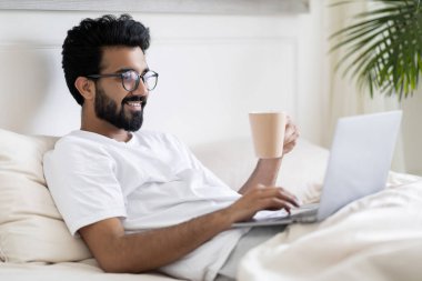 Genç mutlu Hintli adam dizüstü bilgisayar kullanıyor ve yatakta otururken kahve içiyor, yakışıklı doğu çocuğu internete bakıyor, yatak odasında dinleniyor, hafta sonu tatilinin tadını çıkarıyor, boş zamanın tadını çıkarıyor.
