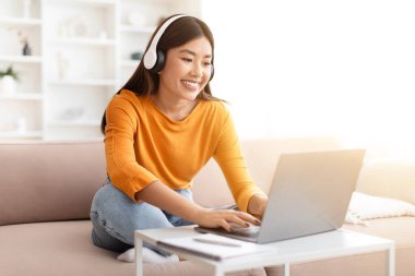 Neşeli gülümseyen güzel Asyalı kadın dizüstü bilgisayar ve kablosuz kulaklık kullanıyor, evdeki koltukta oturuyor, bilgisayar klavyesinde daktilo yazıyor, öğrencinin internet dersi var, webinar, kopyalama alanı var.