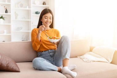 İnce, neşeli, genç Asyalı kadın gündelik kıyafetlerini giymiş, kanepede bacakları havada oturuyor, evde sağlıklı sebze salatası yiyor, öğle yemeğinin, fotokopi alanının, güneş ışığının tadını çıkarıyor. Diyet, beslenme konsepti
