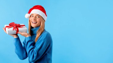 Kazaklı ve Noel Baba şapkalı mutlu bir bayan, elinde hediye kutusuyla boş alana bakıyor, mavi stüdyo arka planında izole edilmiş, panorama. Yeni yıl için harika bir hediye, Noel kutlaması.