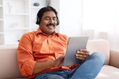 Olumlu, olgun, bıyıklı, dijital tablet ve kablosuz kulaklık kullanarak evdeki koltukta oturan, internet içeriğini izleyen, video görüşlü, müzik dinleyen, yer kopyalayan Hintli adam.