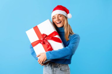 Noel Baba şapkası takan neşeli genç bir kadın, sıcacık bir şekilde Noel hediyesini kucaklıyor, gülümseyişi neşe saçıyor ve mavi stüdyo arka planına karşı mutluluk saçıyor.