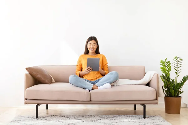 Happy pretty young asian woman sitting on couch, using modern digital tablet at home, chatting with guys on dating app, checking social media, reading blog, copy space for ad, living room interior