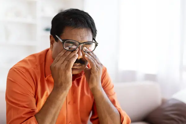 Tired indian mature man rubbing eyes under eyeglasses, sitting on couch at home interior, suffering from sore eyes syndrome, exhausted after difficult day, copy space, closeup. Eyes health, eyesight