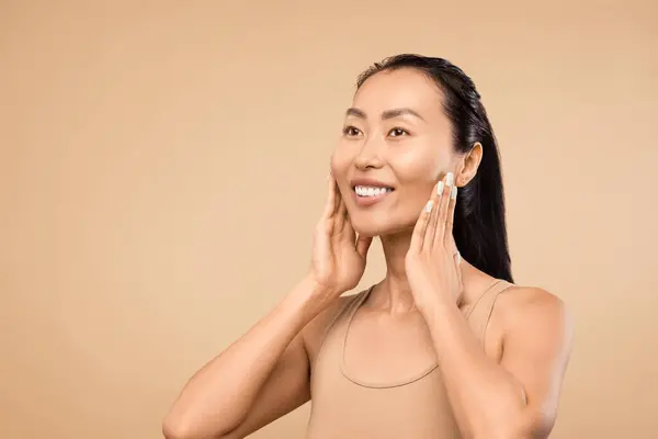 Smiling Asian woman with radiant skin, touches her face, exemplifying skincare results and natural beauty against beige background, free space