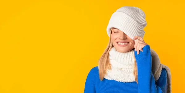 明るい冬のオファー コピースペース付きのパノラマ イエロースタジオの背景にポーズ ホワイトニットの帽子で片目を覆うケアフリー幸せな女性 — ストック写真