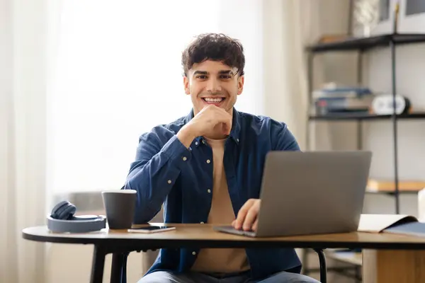 ポジティブなアラブガイウェブサーフィングとカメラに笑顔 ラップトップコンピュータでオンラインで作業 現代のオフィスのインテリアで職場テーブルに座って コンピュータワークとEラーニングコンセプト — ストック写真