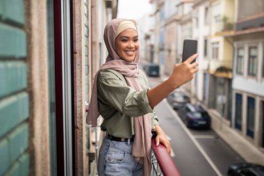 İyi ki tesettürlü genç bir bayan cep telefonuyla selfie çekiyor, apartmanın balkonunda duruyor, akıllı telefon cihazıyla eğleniyor. Sosyal Medya Günlüğü ve Gadget Yaşam Biçimi