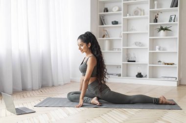 Çevrimiçi Egzersiz Yapan Genç Doğulu Kadın, İç İşleri. Atletik güzel Hintli kadın yoga yapıyor, internetten video izliyor, yan görüş alıyor, uzayı kopyalıyor.